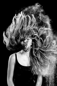 Vrouw zwiept haar haar omhoog door een wolk poeder, gezicht schuil achter het haar, zwart-wit