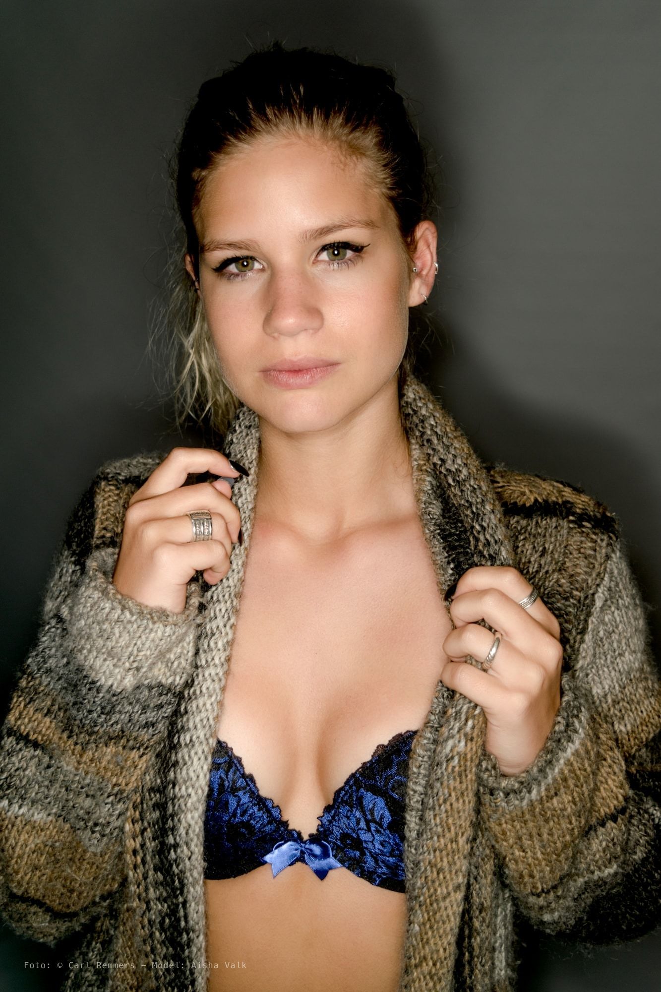 Vrouw in een grofgebreid vest over blauwe kanten lingerie, handen aan de kraag, grijze achtergrond