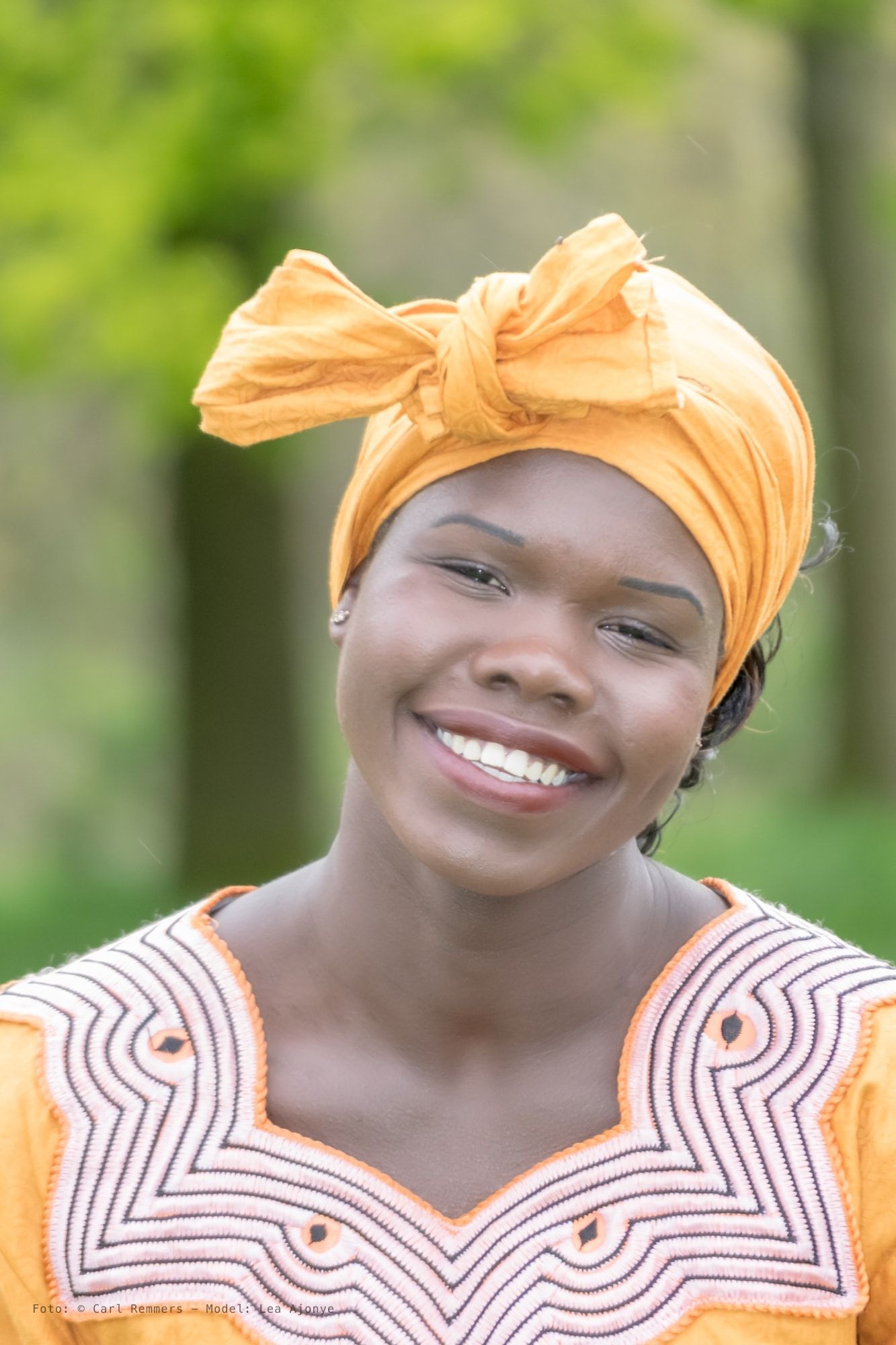Lachende vrouw met een oranje hoofddoek, buiten tussen groene bomen