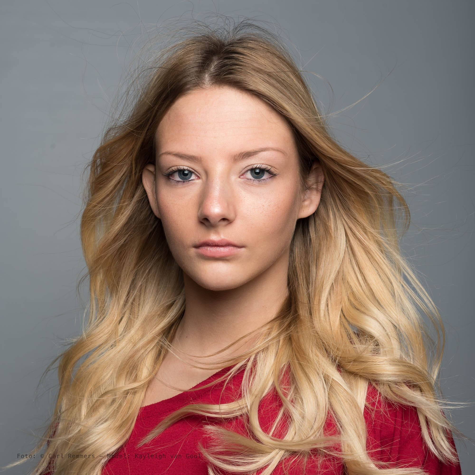 Vrouw met blond golvend haar in een rode top, grijze achtergrond