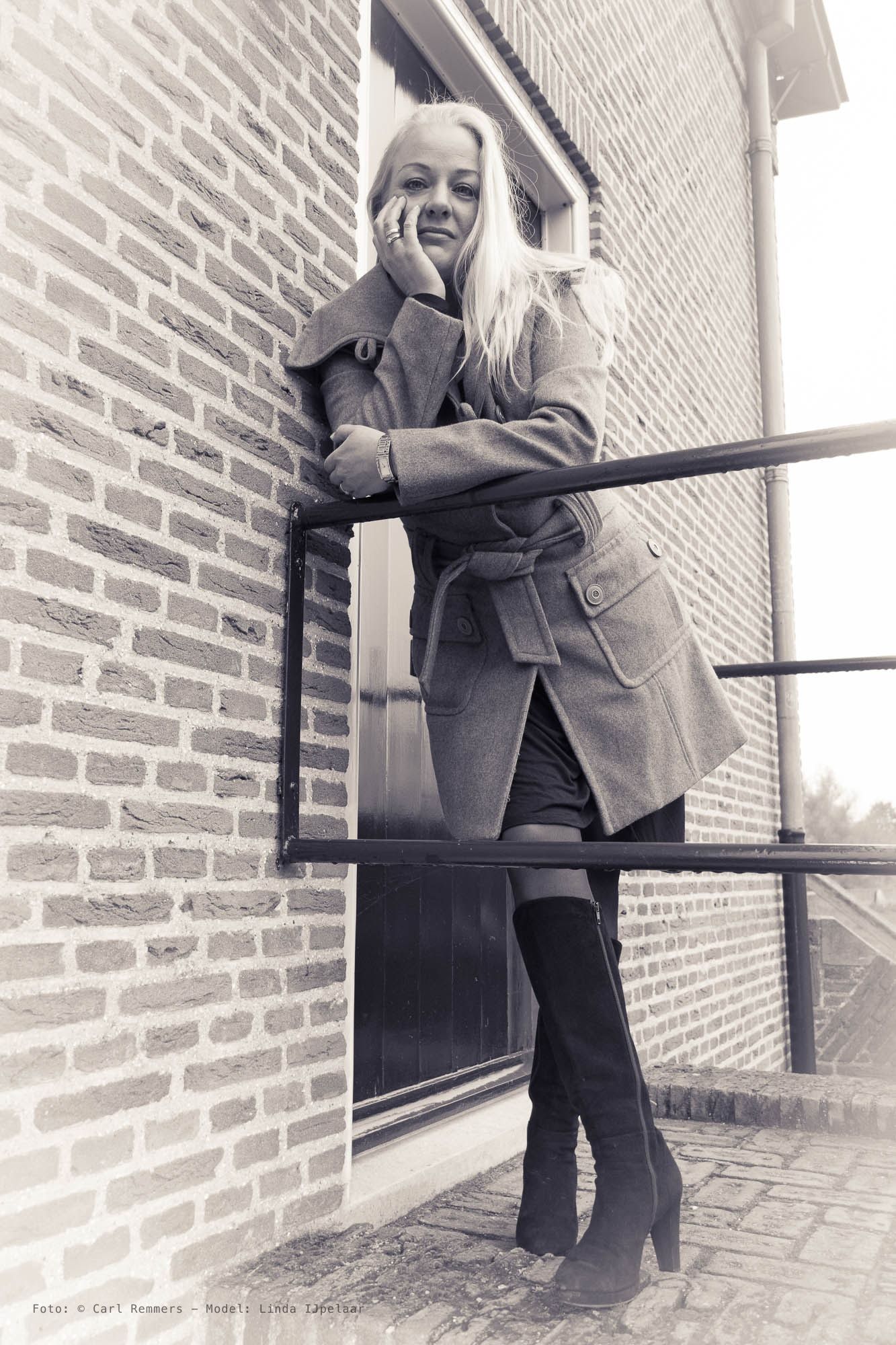 Vrouw in een wollen jas en hoge laarzen leunt over een balustrade, bakstenen muur, sepia