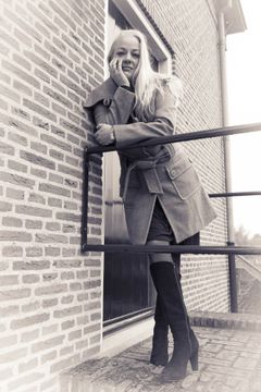 Vrouw in een wollen jas en hoge laarzen leunt over een balustrade, bakstenen muur, sepia