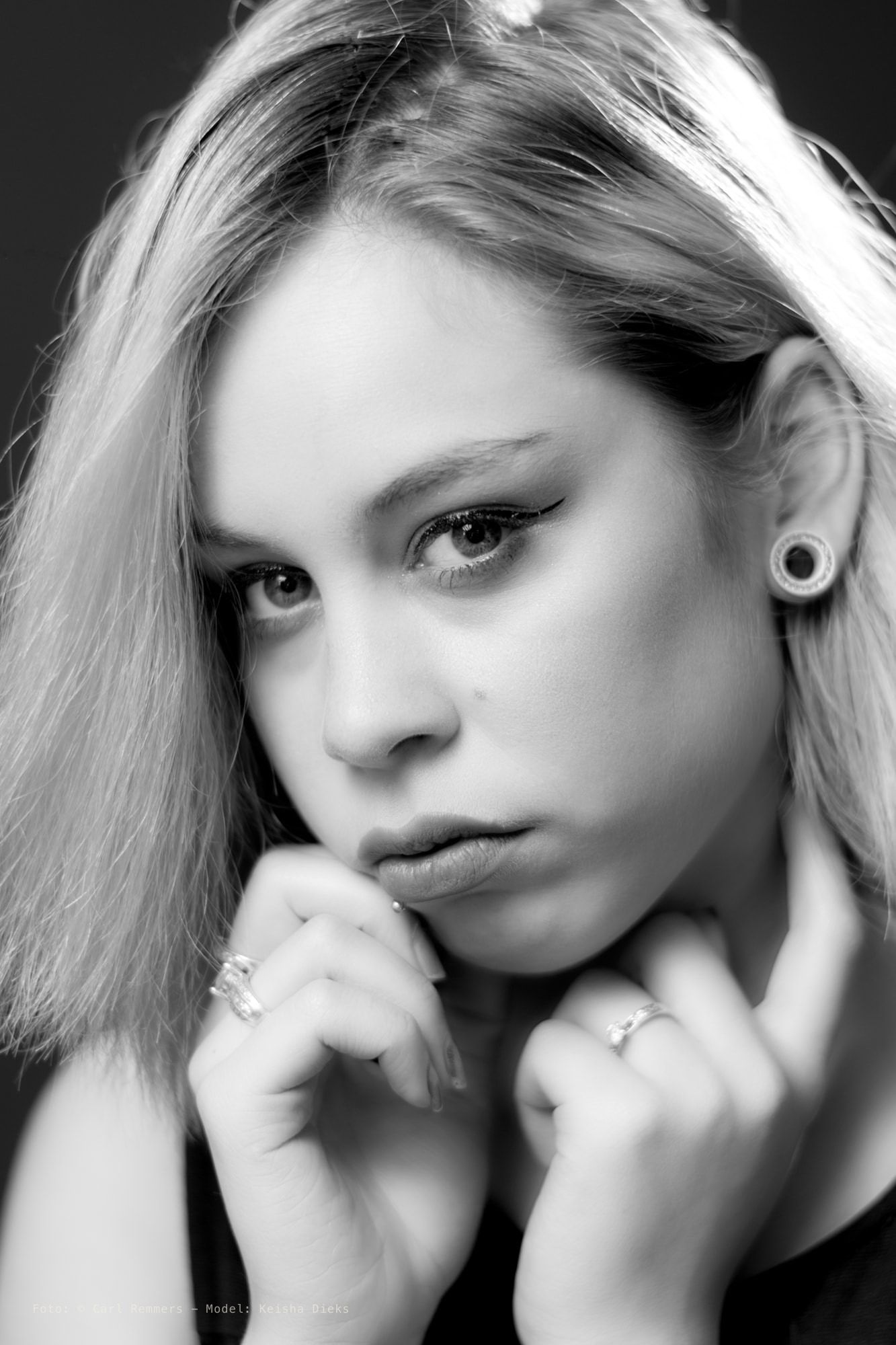 Zwart-witportret van blonde vrouw met beide handen bij kin, ringen en oorplug, donkere achtergrond.