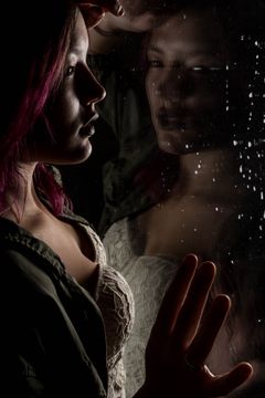 Vrouw met roze haar bij een beslagen raam, haar spiegelbeeld en een hand tegen het glas