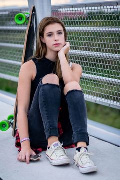 Jonge vrouw zit tegen een lantaarnpaal met een longboard naast zich, buiten op een brug