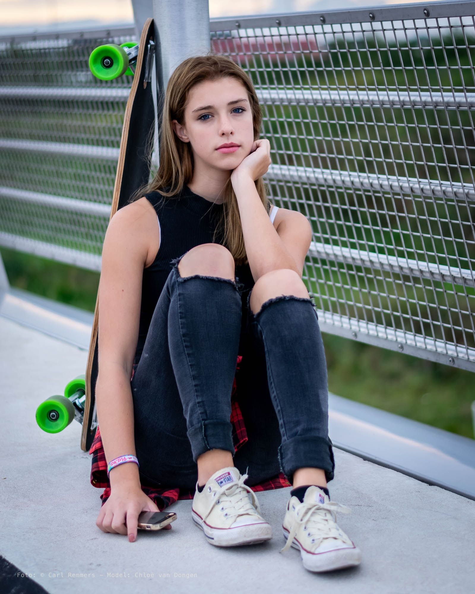 Young woman sitting against a lamp post with a longboard beside her, outside on a bridge