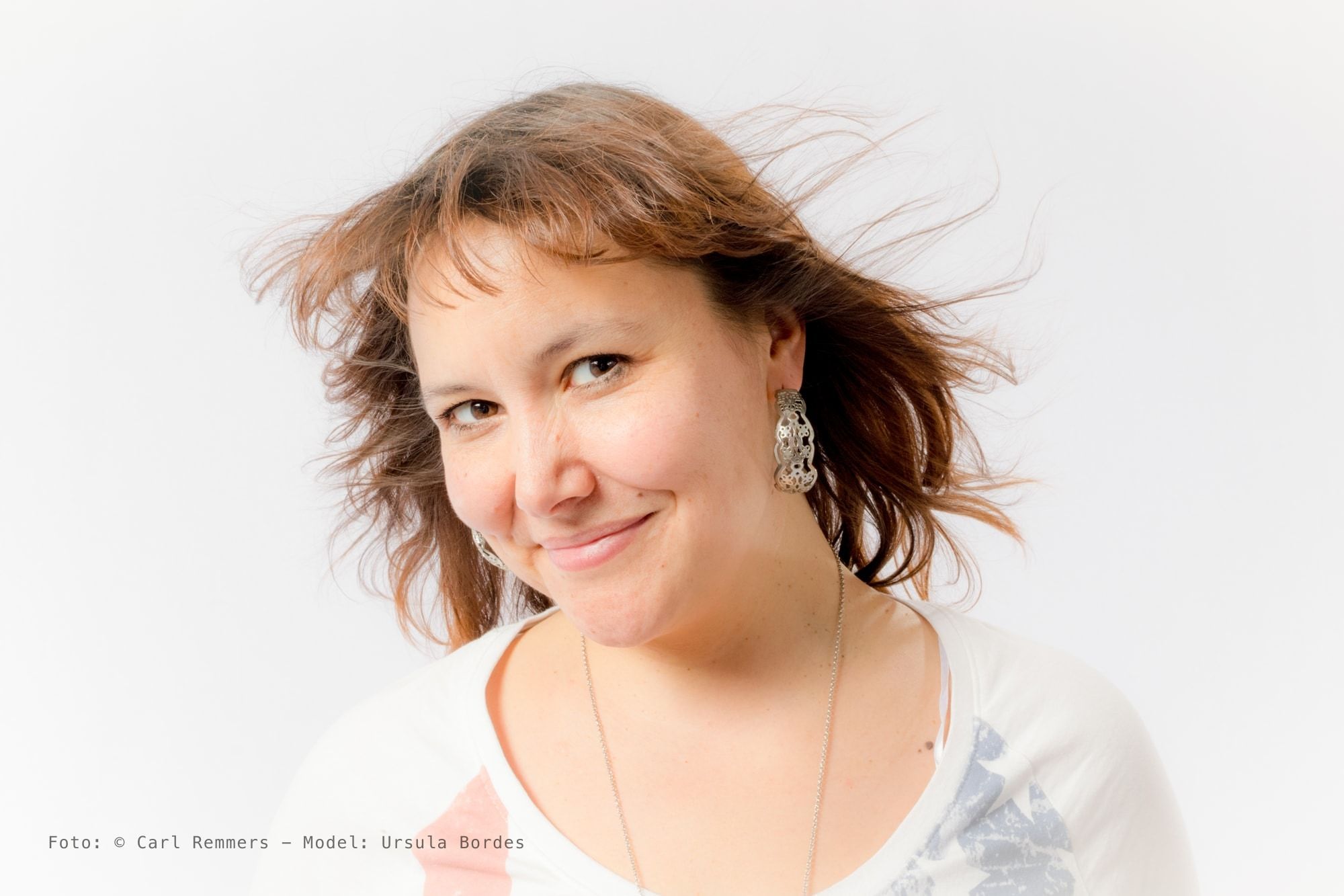 Laughing woman with brown hair blowing up, white printed shirt, fine necklace, against a white background.