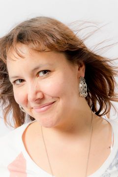 Lachende vrouw met opwaaiend bruin haar, witte shirt met print, fijne halsketting, tegen witte achtergrond.